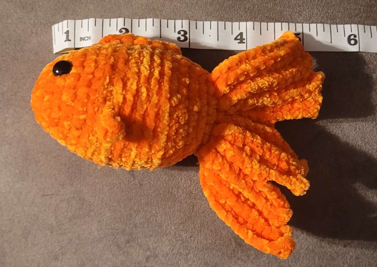 Bright orange crocheted goldfish plush with flowing fins, shown with measuring tape